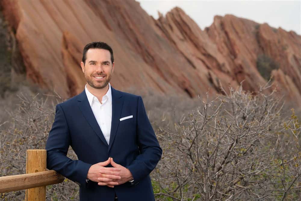 Brendan McQuaid in a navy suit at a wooden fence, red sandstone formations behind him